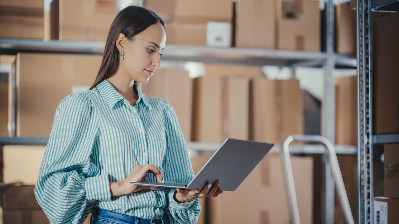Portrait of a Worker Checking Inventory, Writing Down Notes on Laptop Computer. Young Woman Working in a Warehouse Storeroom with Rows of Shelves with Parcels, Packages with Orders Ready for Shipment.