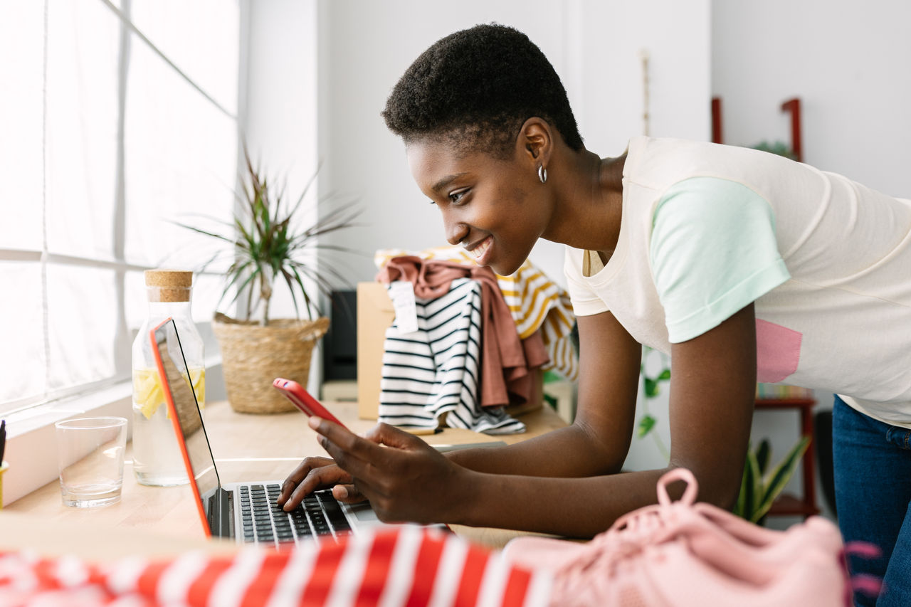 Small business owner woman looking at phone and laptop in the middle of her clothing inventory