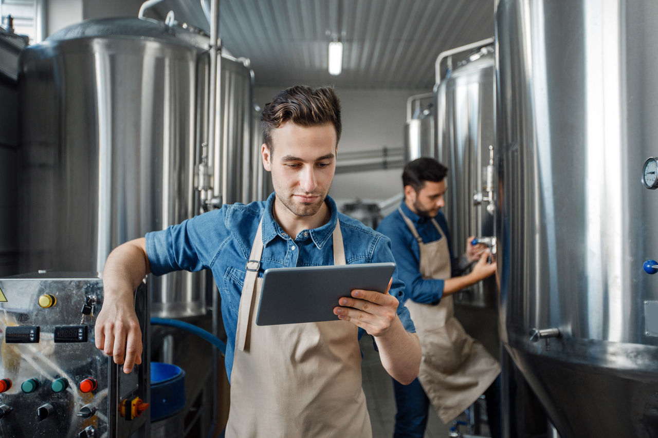 Smart factory, modern brewery management and craft beverage production. Busy young handsome guys workers in aprons, use tablet for operate kettle, steel vats in plant interior, copy space