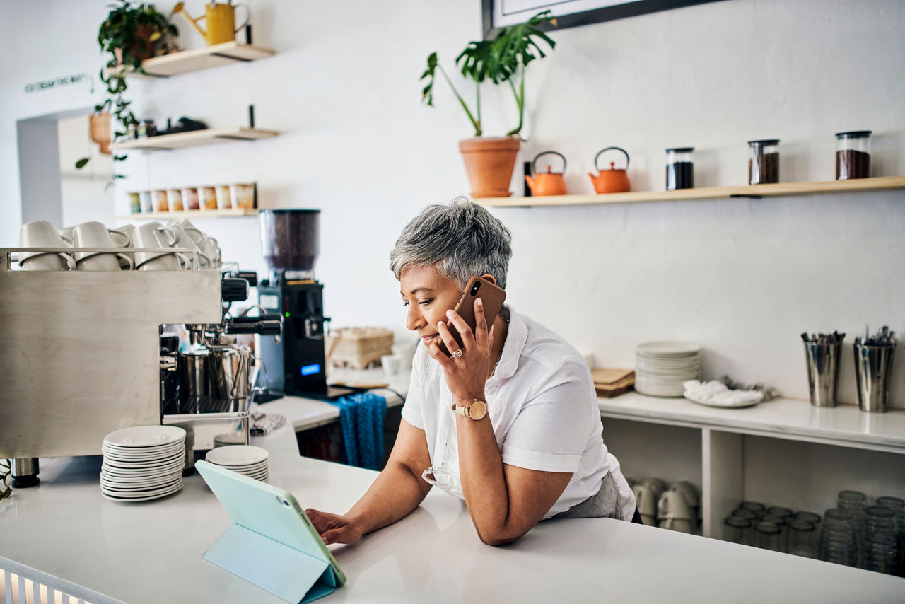 Old woman in coffee shop, small business with tablet and phone call, digital invoice and chat with supplier and barista. Technology, inventory check and communication with wifi and manage orders
