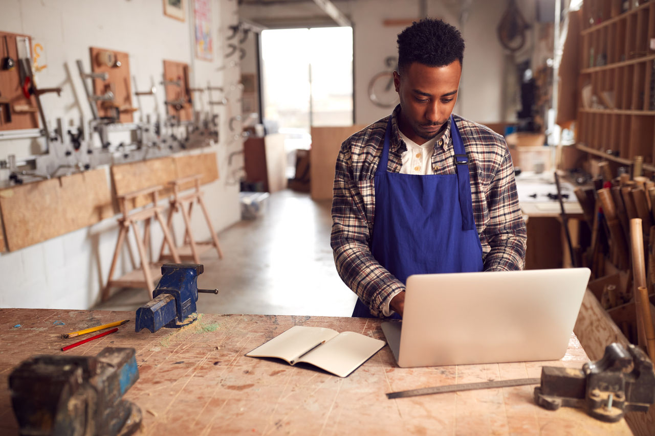 Male Craftsman In Carpentry Workshop On Laptop
