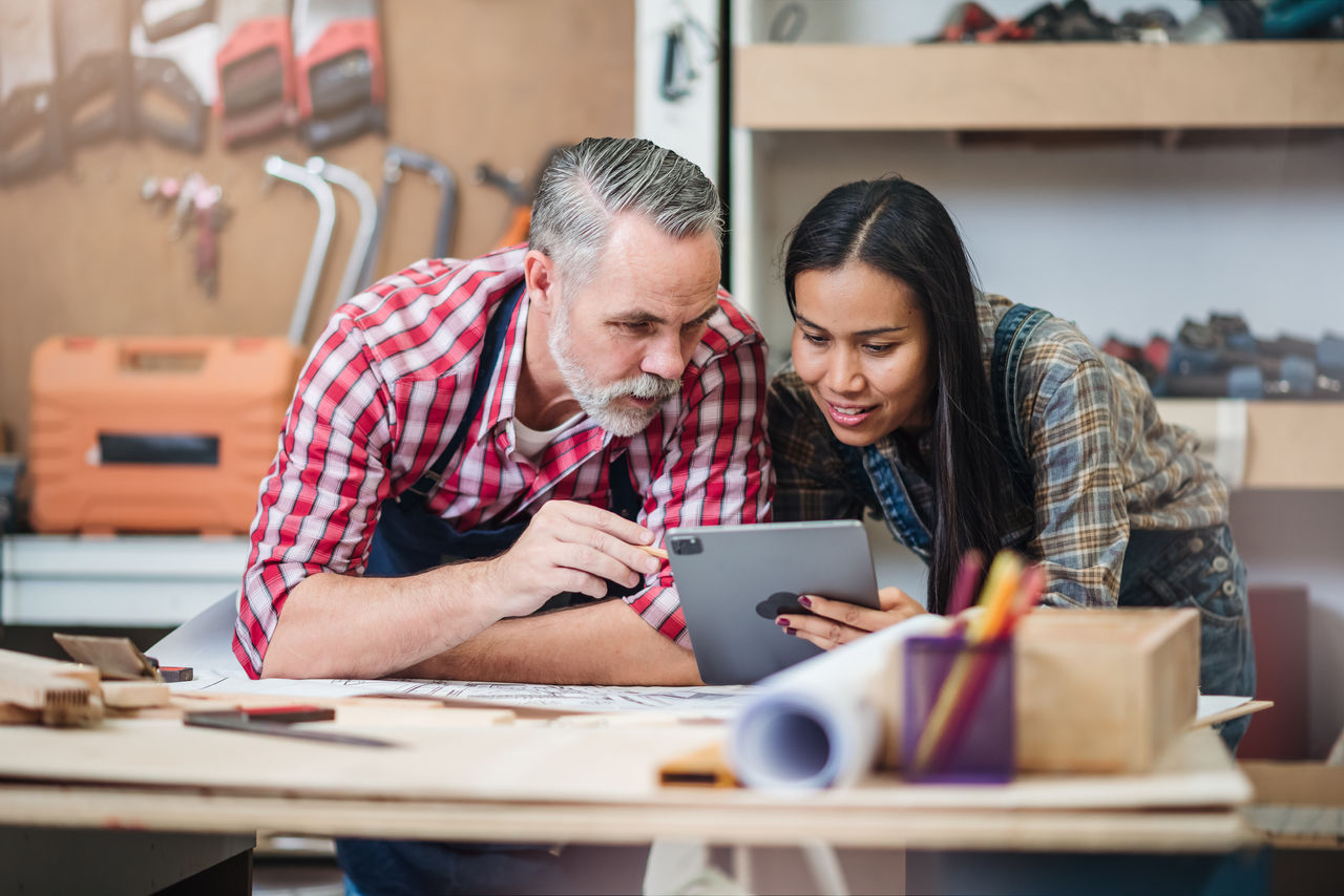 Small business furniture owner and professional architect interior designer working in carpenter workshop with blueprint and tablet discussing about home furnishing design project.