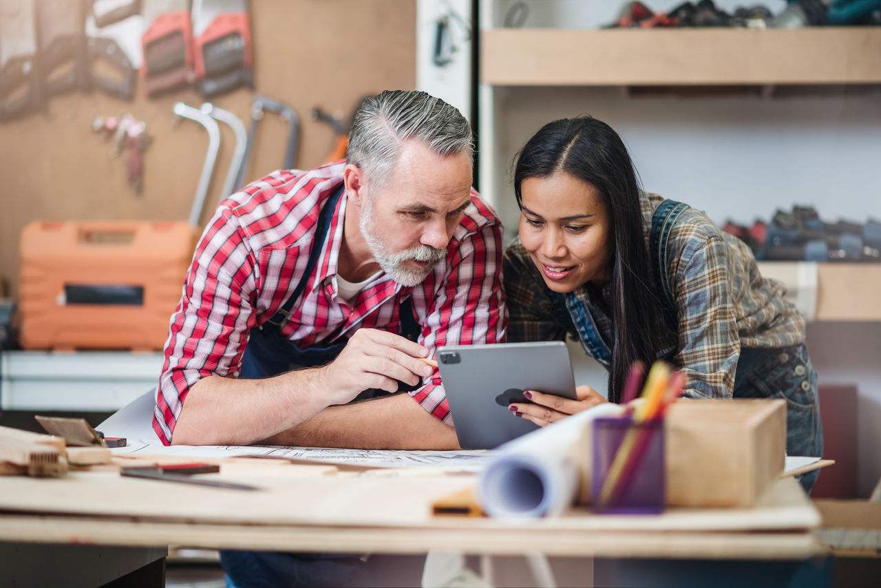 Small business furniture owner and professional architect interior designer working in carpenter workshop with blueprint and tablet discussing about home furnishing design project.