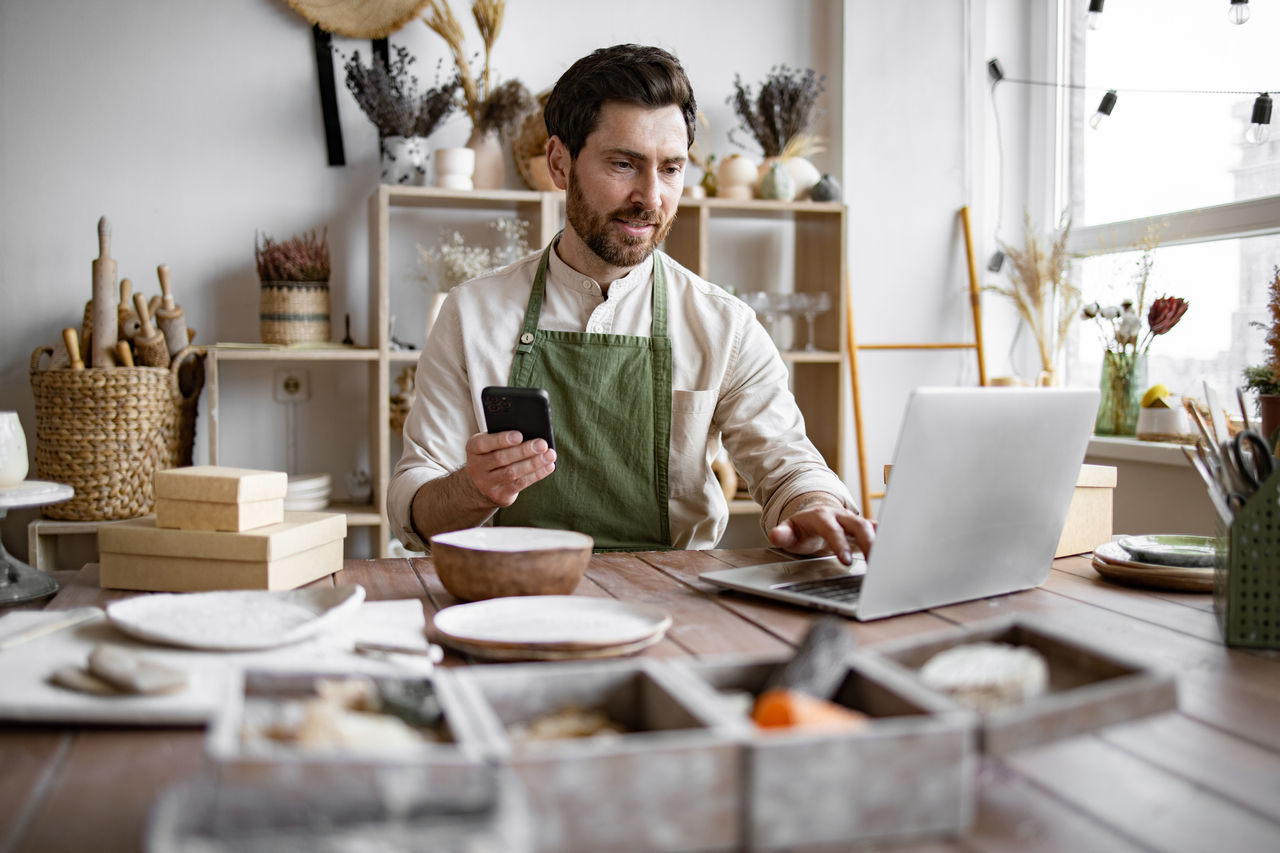 Successful guy store employee with ceramic tableware shoots photos on smartphone. Charismatic Caucasian man seller in souvenir shop takes pictures of goods at phone for posting on social networks.