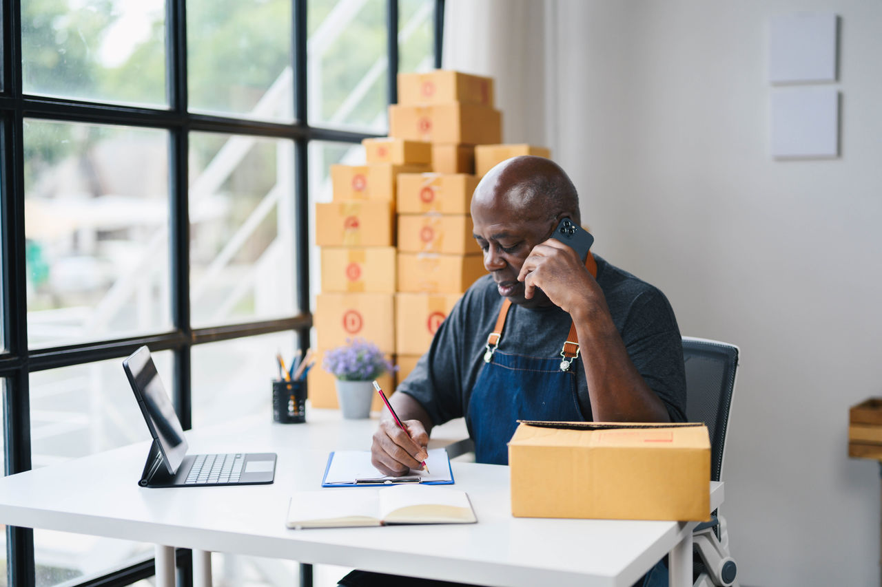 Man managing online business orders from home office, talking on phone, writing notes, surrounded by cardboard boxes.